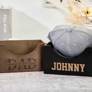 Könnte beinhalten: Zwei hölzerne Hutboxen mit personalisierten Namen. Die Box auf der linken Seite ist braun und trägt die Aufschrift "DAD" mit den Namen "clare", "monica" und "jessie" darunter. Die Box auf der rechten Seite ist schwarz und trägt die Aufschrift "JOHNNY" in goldenen Buchstaben.