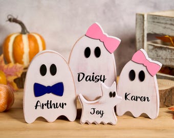 Può includere: Decorazioni a forma di fantasma in legno con occhi neri e nomi: Arthur, Daisy, Karen e Joy. I fantasmi hanno una finitura bianca invecchiata. Daisy e Karen hanno fiocchi rosa. Arthur ha un papillon blu. Zucca e foglie d'autunno sullo sfondo.