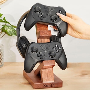 May include: A wooden stand with a personalised name "Smith" engraved on it. The stand holds two black video game controllers and a pair of black headphones.