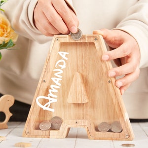 Puede incluir: Una alcanc&iacute;a de madera con forma de la letra A con el nombre "Amanda" escrito en ella. Una mano est&aacute; poniendo una moneda en la ranura.