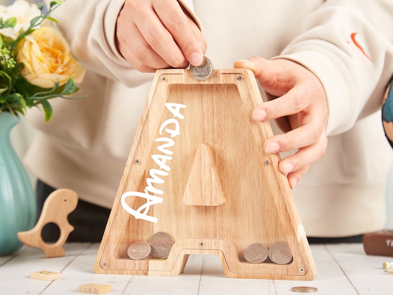 Puede incluir: Una alcanc&iacute;a de madera con forma de la letra A con el nombre "Amanda" escrito en ella. Una mano est&aacute; poniendo una moneda en la ranura.