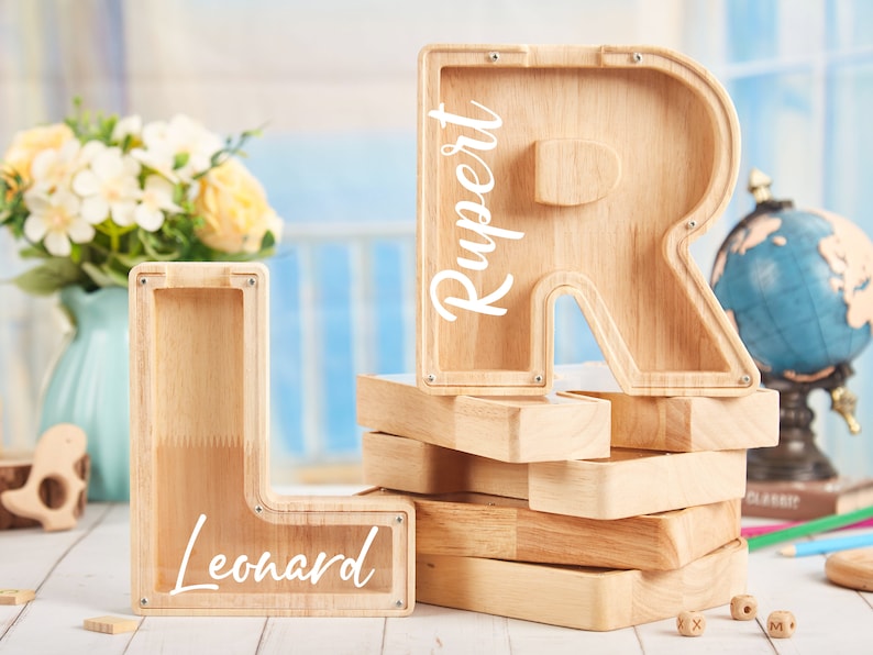 Puede incluir: Dos alcanc&iacute;as de madera en forma de letras, una con forma de L y la otra con forma de R. La alcanc&iacute;a L tiene el nombre "Leonard" escrito en ella y la alcanc&iacute;a R tiene el nombre "Rupert" escrito en ella.