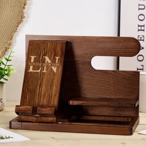 May include: A dark brown wooden phone stand with a phone holder, a watch holder, and a small shelf. The stand has the initials "LN" and the text "LOUISE NEWTON" engraved on the front.