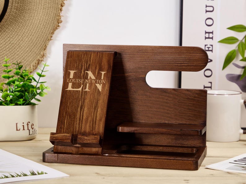 May include: A dark brown wooden phone stand with a phone holder, a watch holder, and a small shelf. The stand has the initials "LN" and the text "LOUISE NEWTON" engraved on the front.