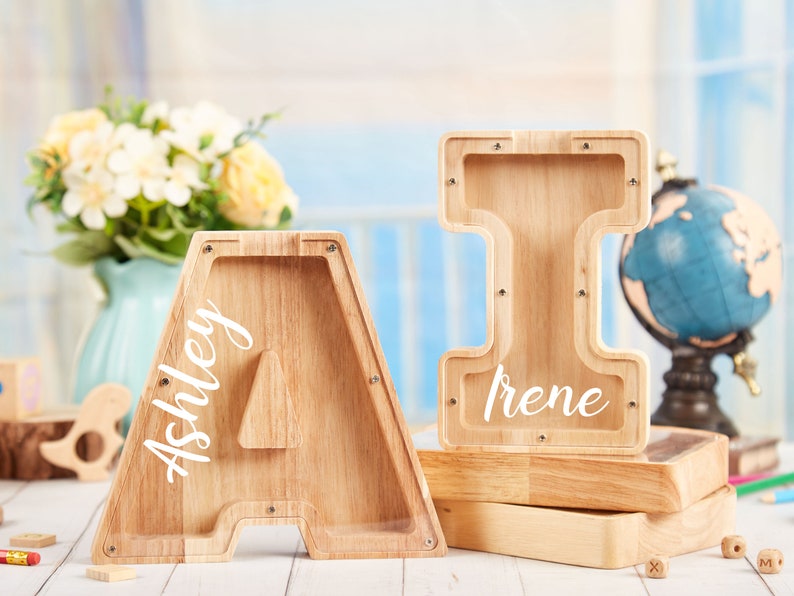 Puede incluir: Dos alcanc&iacute;as de madera con forma de letras, una es la letra A con el nombre Ashley escrito en ella, la otra es la letra I con el nombre Irene escrito en ella.