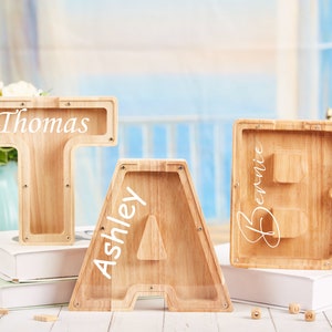 Puede incluir: Tres huchas de madera en forma de letras, cada una con un nombre inscrito en la parte delantera. Las letras son T, A y B, y los nombres son Thomas, Ashley y Bernie.