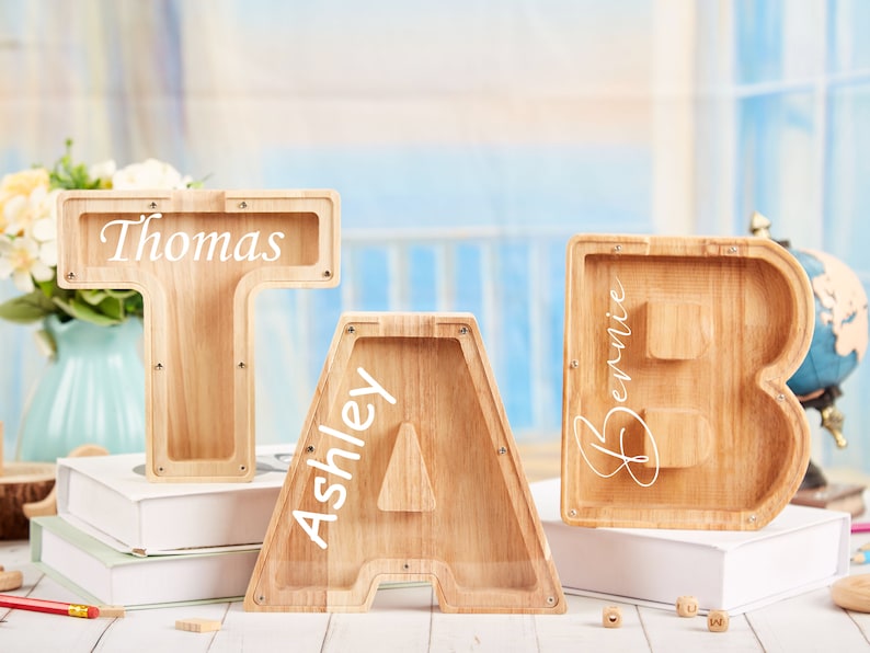 Puede incluir: Tres huchas de madera en forma de letras, cada una con un nombre inscrito en la parte delantera. Las letras son T, A y B, y los nombres son Thomas, Ashley y Bernie.