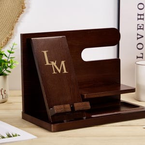 May include: A dark brown wooden organiser with a phone stand, a watch holder, and a tray. The phone stand has a slot for a phone and a small shelf for charging cables. The watch holder has a small shelf for a watch and a slot for a watch band. The tray is a small, rectangular tray for holding keys, coins, or other small items. The organiser has the initials "L.M." engraved on the front.