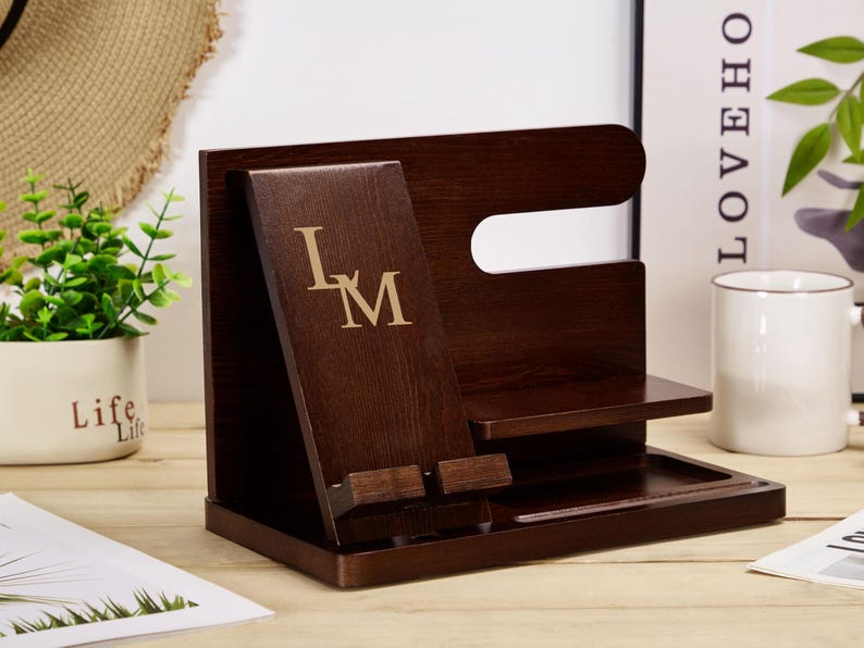 May include: A dark brown wooden organiser with a phone stand, a watch holder, and a tray. The phone stand has a slot for a phone and a small shelf for charging cables. The watch holder has a small shelf for a watch and a slot for a watch band. The tray is a small, rectangular tray for holding keys, coins, or other small items. The organiser has the initials "L.M." engraved on the front.