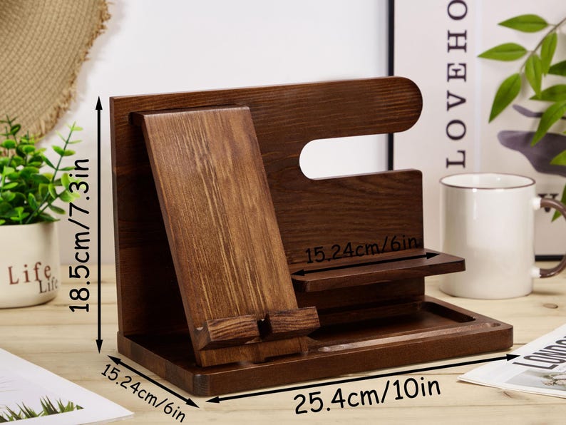 May include: A wooden phone stand with a slot for a phone, a small shelf, and a tray. The stand is 25.4 cm wide, 15.24 cm deep, and 18.5 cm tall.