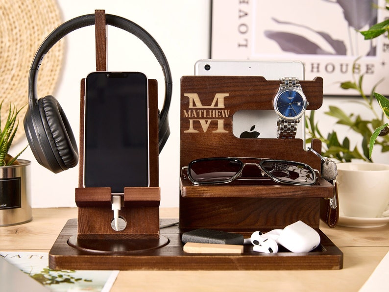 May include: A wooden organizer with a personalized name, "Matthew", engraved on the front. It features slots for a phone, watch, sunglasses, and headphones. There is also a small compartment for earbuds or other small items.