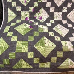 May include: A handmade quilt with a geometric pattern. The quilt features squares, diamonds, and triangles in shades of green, purple, and white against a dark gray background. The quilt is bordered with a white edge.