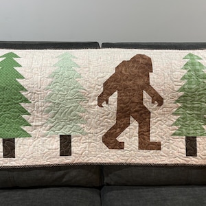 May include: A Bigfoot-themed quilt featuring a brown silhouette of the creature walking between green and light green trees. The quilt has a cream background with a brown border, perfect for a cabin or nature-themed decor.