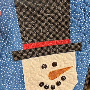 May include: A quilted snowman wall hanging with a black and white gingham hat, red scarf, and white face with black buttons for eyes and a carrot nose.