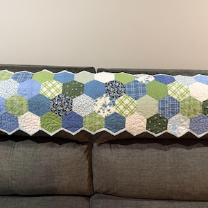 May include: A rectangular quilt featuring a repeating hexagonal patchwork pattern in various shades of blue, green, and white. The fabric pieces display diverse floral, plaid, and solid textures, all finished with a white scalloped edge border.