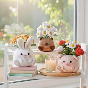 May include: A shelf displays three crochet flower pots: a white bunny, a brown bear, and a pink pig. Each pot holds colorful crochet flowers. A lit candle sits on a wooden tray, adding a warm ambiance to the scene. The shelf is white.