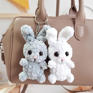 Może przedstawiać: Dwa pluszowe breloczki do kluczy w kształcie króliczków, jeden jasnoniebieski i jeden biały, wiszą na srebrnym łańcuszku przymocowanym do brązowej torebki. Króliczki mają czarne oczy i różowe nosy. Breloczki wykonane są z miękkiego, teksturowanego materiału.