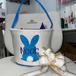 Personalized with buyers choice of name. White canvas Easter Basket with Blue Bunny applique, includes tissue paper, bow and clear gift wrapper