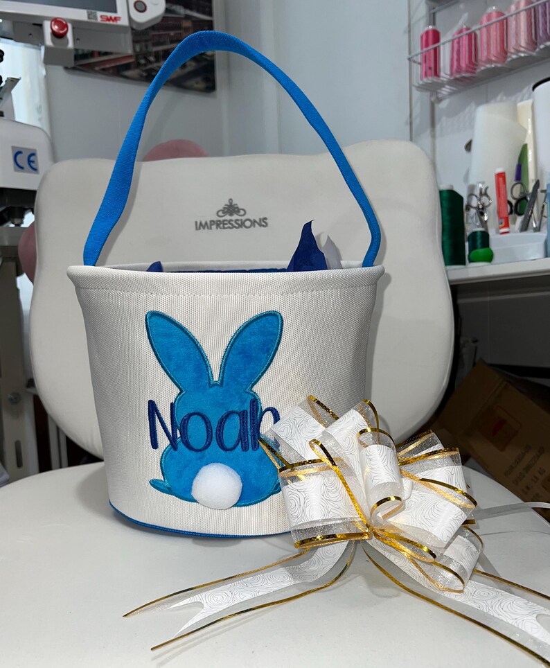 Personalized with buyers choice of name. White canvas Easter Basket with Blue Bunny applique, includes tissue paper, bow and clear gift wrapper