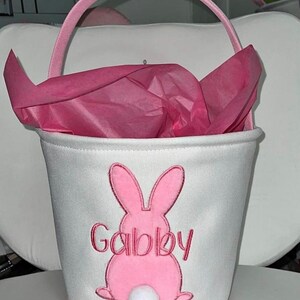 May include: White fabric Easter basket with a pink bunny design and the name "Gabby" embroidered in pink. The basket has a pink handle and is filled with pink tissue paper. A white pom-pom tail is attached to the bunny.