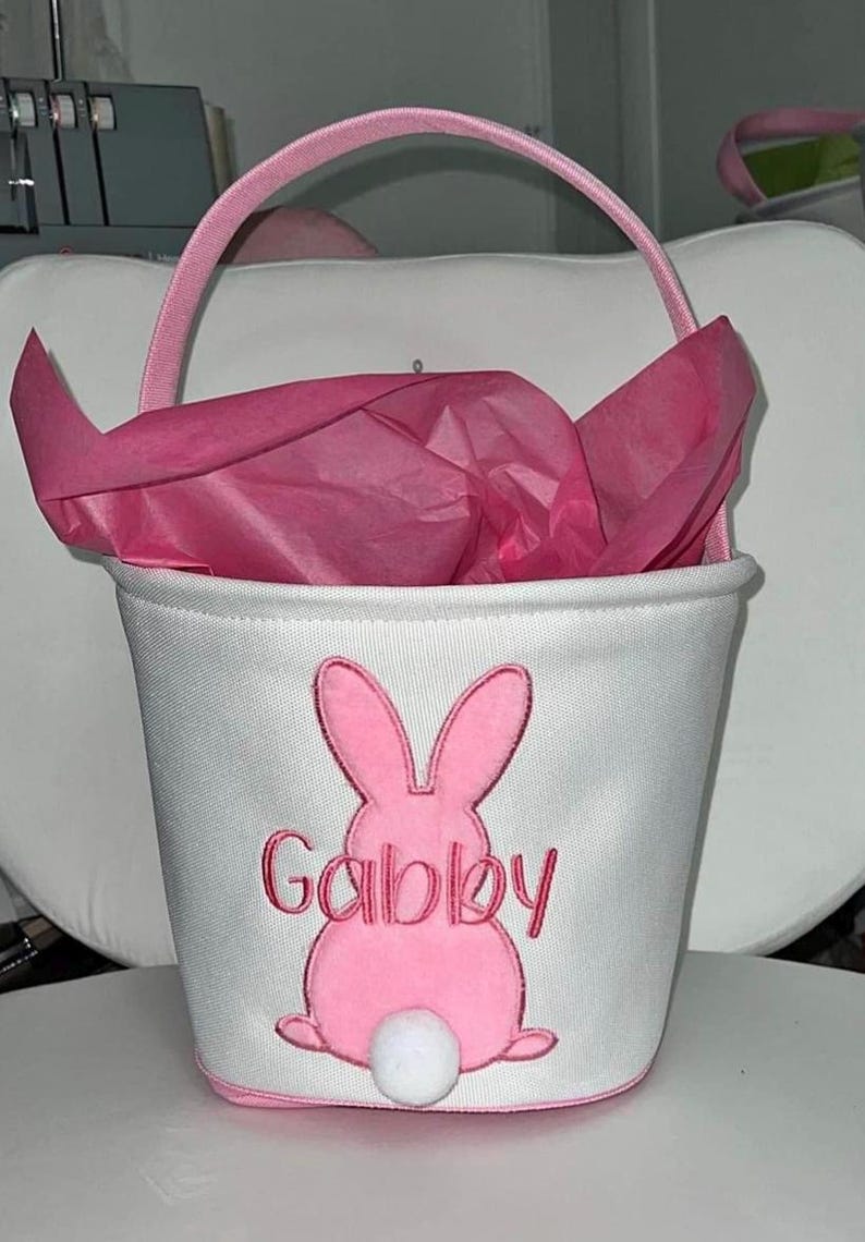 May include: White fabric Easter basket with a pink bunny design and the name "Gabby" embroidered in pink. The basket has a pink handle and is filled with pink tissue paper. A white pom-pom tail is attached to the bunny.