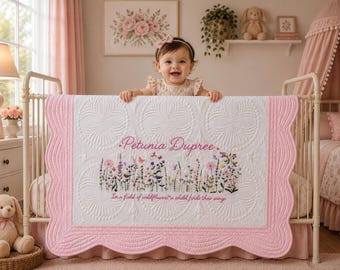 May include: A white quilt with a scalloped pink border, featuring an embroidered wildflower meadow, the name Petunia Dupree, and the text In a field of wildflowers, a child finds their wings. The quilt has a circular quilted pattern throughout.