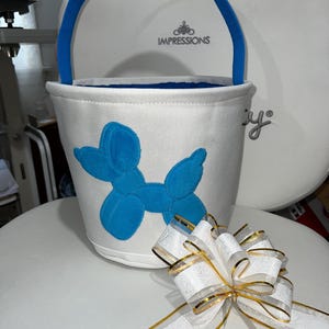 Personalized with buyers choice of name. White canvas Easter Basket with Blue Balloon Dog applique, includes tissue paper, bow and clear gift wrapper. Personalization in blue