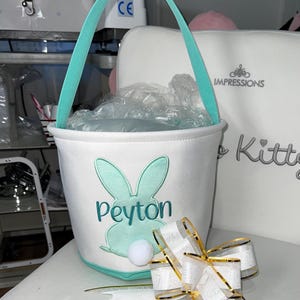 May include: White Easter basket with a teal handle and bunny design. The bunny has teal ears and a white pom-pom tail. The name "Peyton" is embroidered in teal. A white and gold ribbon bow is in the foreground.