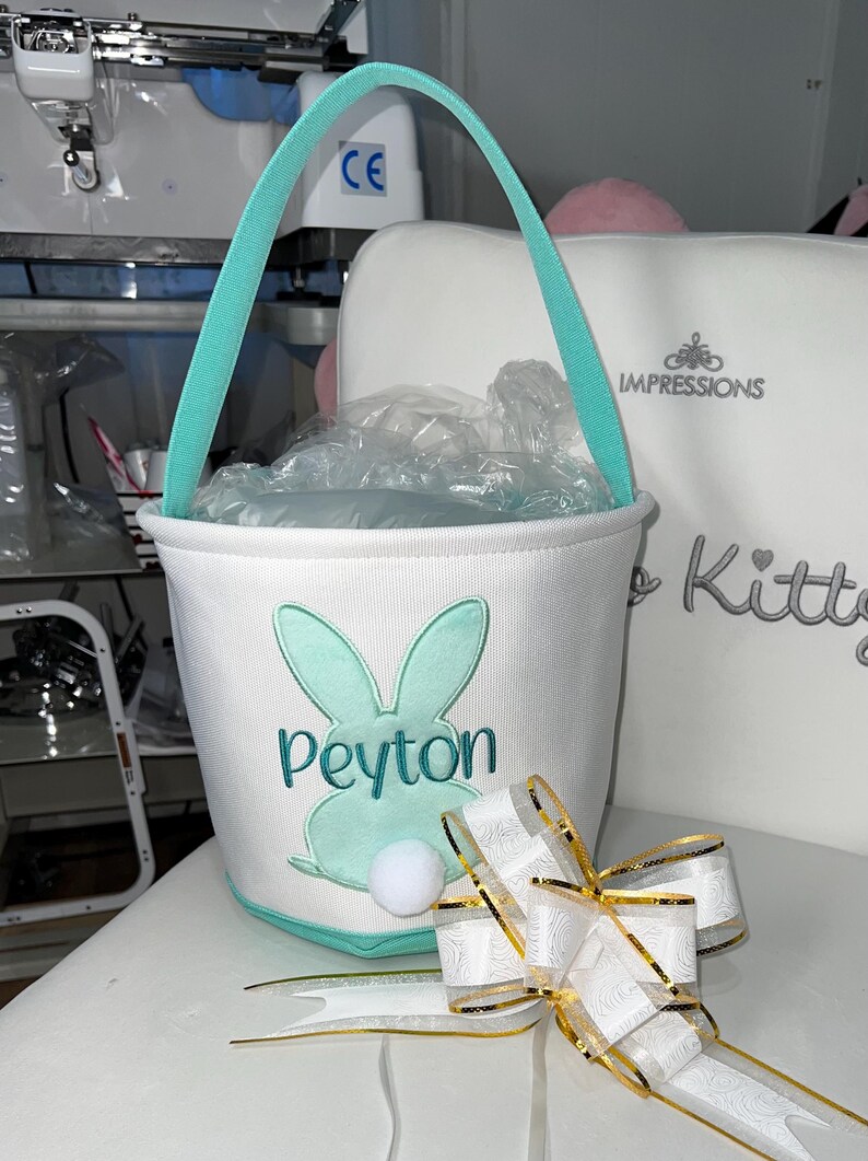 May include: White Easter basket with a teal handle and bunny design. The bunny has teal ears and a white pom-pom tail. The name "Peyton" is embroidered in teal. A white and gold ribbon bow is in the foreground.