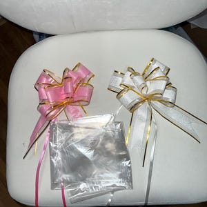 May include: Two decorative bows, one pink and one white, with gold trim and long ribbons. Clear cellophane bags are placed between the bows. The bows are on a white surface, with the words "Hello Kitty" embroidered above.