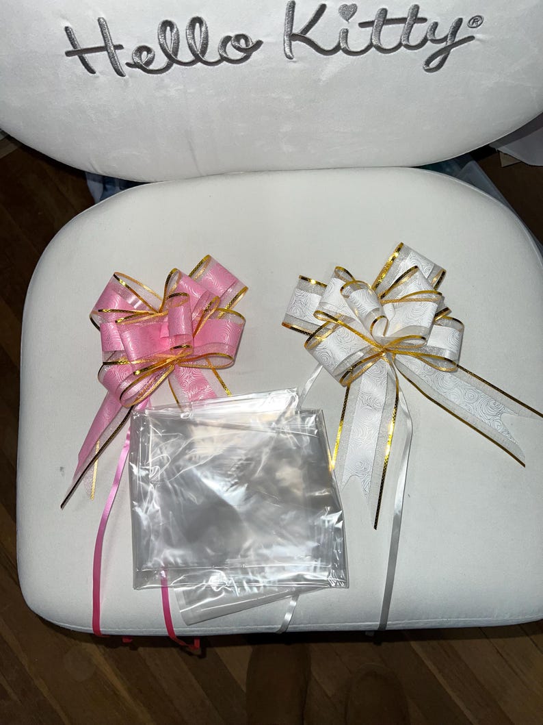 May include: Two decorative bows, one pink and one white, with gold trim and long ribbons. Clear cellophane bags are placed between the bows. The bows are on a white surface, with the words "Hello Kitty" embroidered above.