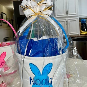 Personalized with buyers choice of name. White canvas Easter Basket with Blue Bunny applique, tissue paper, bow and clear gift wrapper