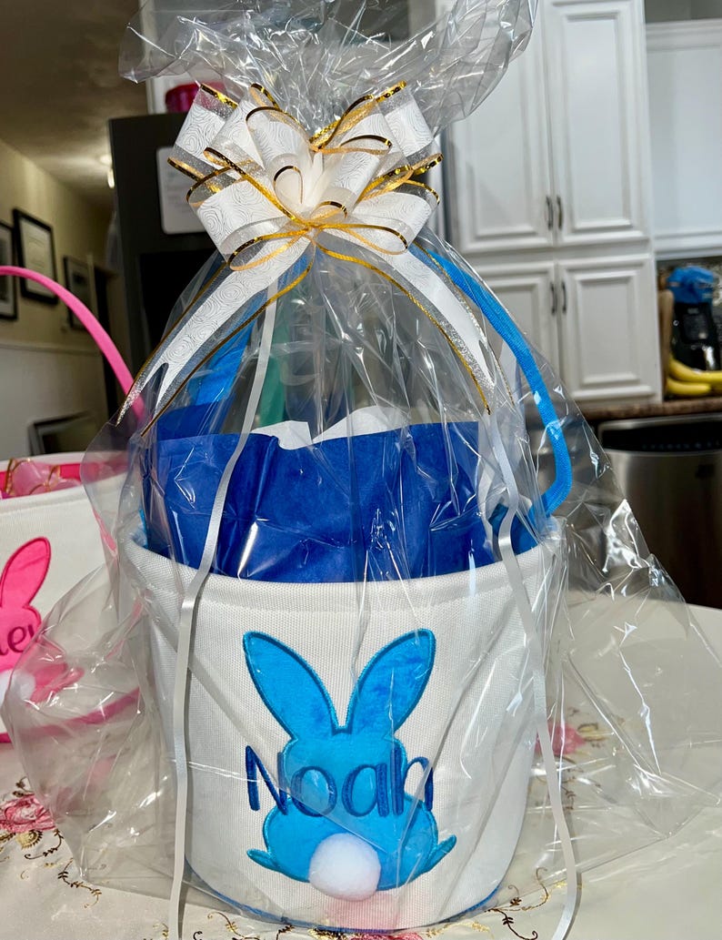 Personalized with buyers choice of name. White canvas Easter Basket with Blue Bunny applique, tissue paper, bow and clear gift wrapper