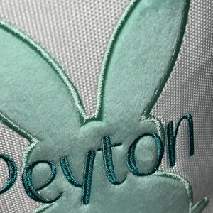 May include: Close-up of a light green bunny design with the name "Peyton" embroidered in teal thread. The bunny has a fluffy texture and a white pom-pom tail. The background is a light gray fabric.