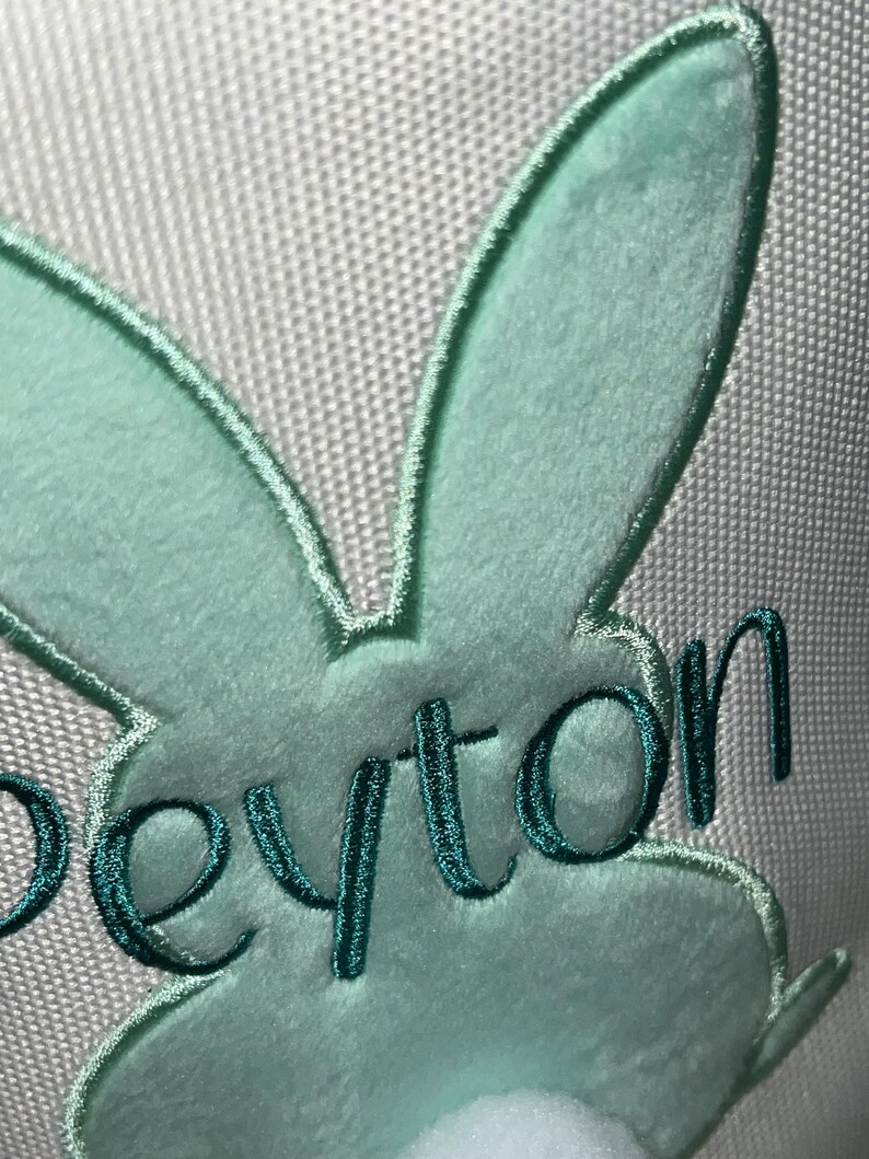 May include: Close-up of a light green bunny design with the name "Peyton" embroidered in teal thread. The bunny has a fluffy texture and a white pom-pom tail. The background is a light gray fabric.