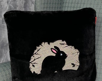 May include: Black plush pillow with a silhouette of a rabbit in a forest scene. The rabbit is black with pink ears and eyes.