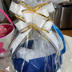May include: A white basket wrapped in clear cellophane, featuring a large decorative bow with white and silver rose patterns and gold metallic edges. The basket contains blue and white tissue paper, topped with a blue handle.