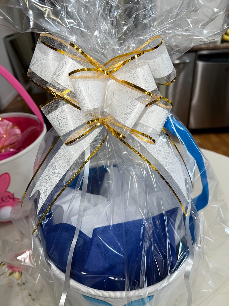 May include: A white basket wrapped in clear cellophane, featuring a large decorative bow with white and silver rose patterns and gold metallic edges. The basket contains blue and white tissue paper, topped with a blue handle.