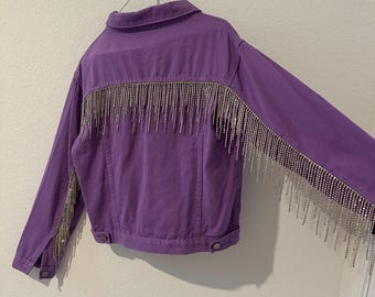 May include: A purple denim jacket with long sleeves and silver rhinestone fringe detailing across the back and sleeves. The jacket has a classic collar and button closures.