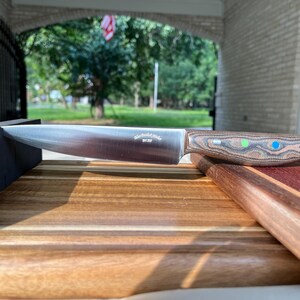 May include: A stainless steel kitchen knife with a wood handle. The handle has green and blue accents. The knife is resting on a wooden cutting board.