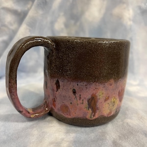 May include: A handmade ceramic mug with a dark brown exterior and a pink and gold glazed lower half. The mug has a curved handle and a textured surface, perfect for enjoying coffee or tea.