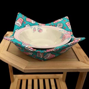 May include: A fabric bowl cosy featuring a teal background with a repeating pattern of coffee cups and small red hearts. Inside the cosy sits a round, cream-coloured ceramic bowl with a light pink rim and a delicate floral design of pink roses.