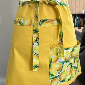 May include: A yellow apron with a lemon print. The apron has a large pocket on the right side and a tie at the waist.