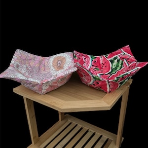 May include: Two fabric bowl cozies. One is pink with a floral pattern, and the other features a watermelon design. They are displayed on a light brown wooden side table against a black background.