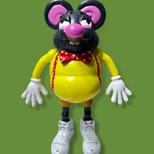 Puede incluir: Una figurita de ratón negro y amarillo con una pajarita roja y zapatillas blancas. El ratón tiene una nariz rosa y orejas grandes.