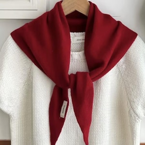 May include: A white short-sleeved knit sweater with a crew neckline and a solid red knit scarf tied around the neck. The sweater features a visible interior label reading about mo, and the red scarf has a small white tag labeled 100% wool.
