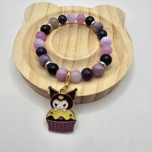 May include: A purple, pink, and black beaded bracelet with a charm of a black cat in a cupcake. The bracelet is on a wooden surface.