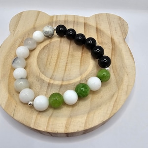May include: A bracelet made with white, green, and black beads. The bracelet is on a wooden surface.