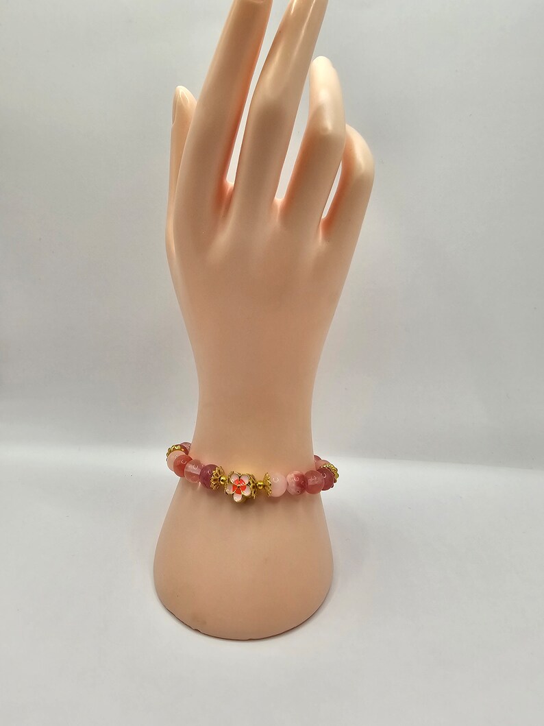Crystal bracelet made of strawberry quartz, peach rice topaz, watermelon quartz and red chalcedony with gold flower and ball spacers and large gold sakura flower charm detailed with pink enamel.