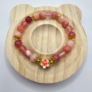 Crystal bracelet made of strawberry quartz, peach rice topaz, watermelon quartz and red chalcedony with gold flower and ball spacers and large gold sakura flower charm detailed with pink enamel.